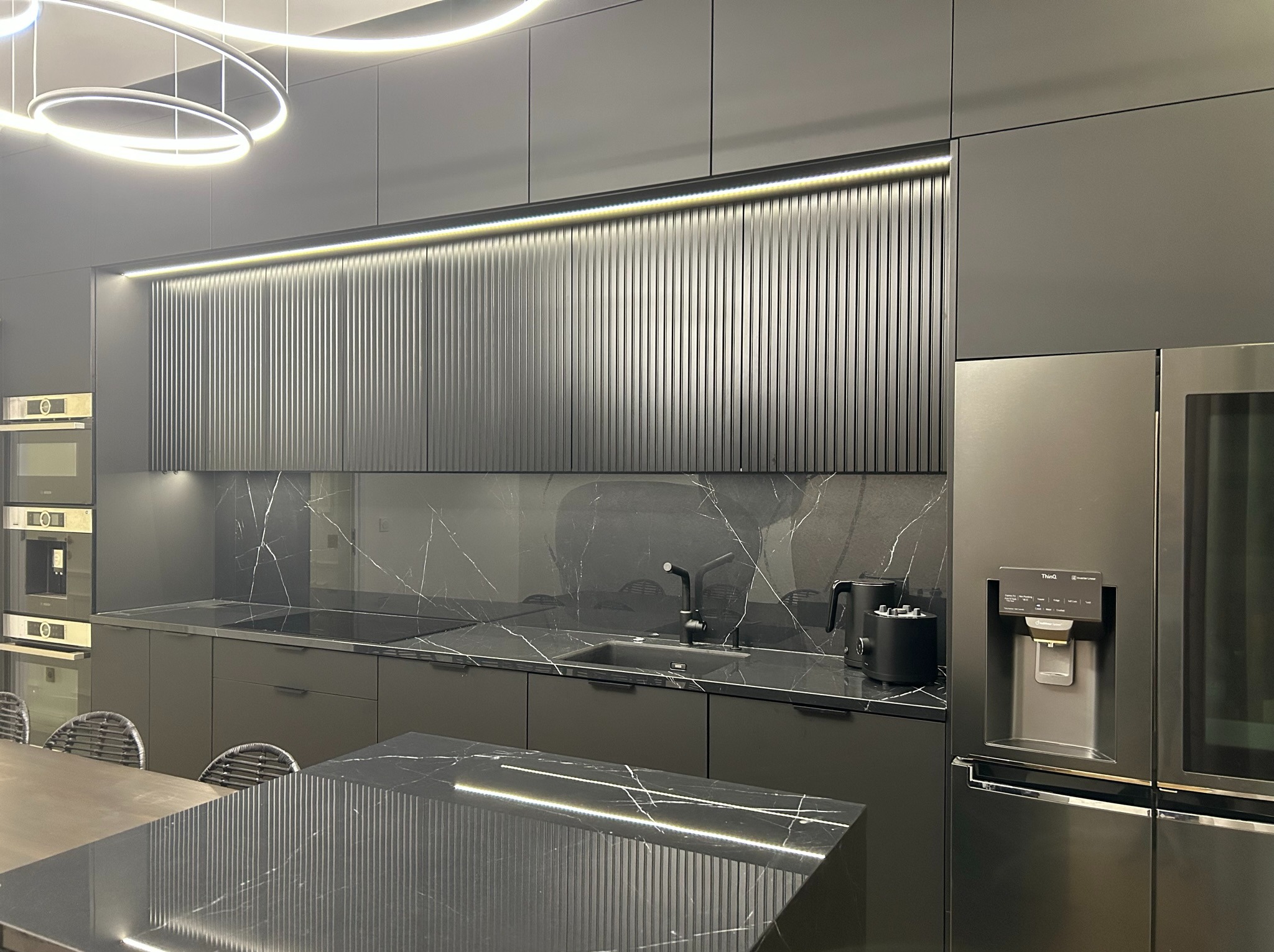 Nowoczesna kuchnia w odcieniach szarości z czarnymi blatami marmurkowymi, podświetlanymi szafkami górnymi z pionowymi żłobieniami oraz wyspą z dodatkowym oświetleniem LED.