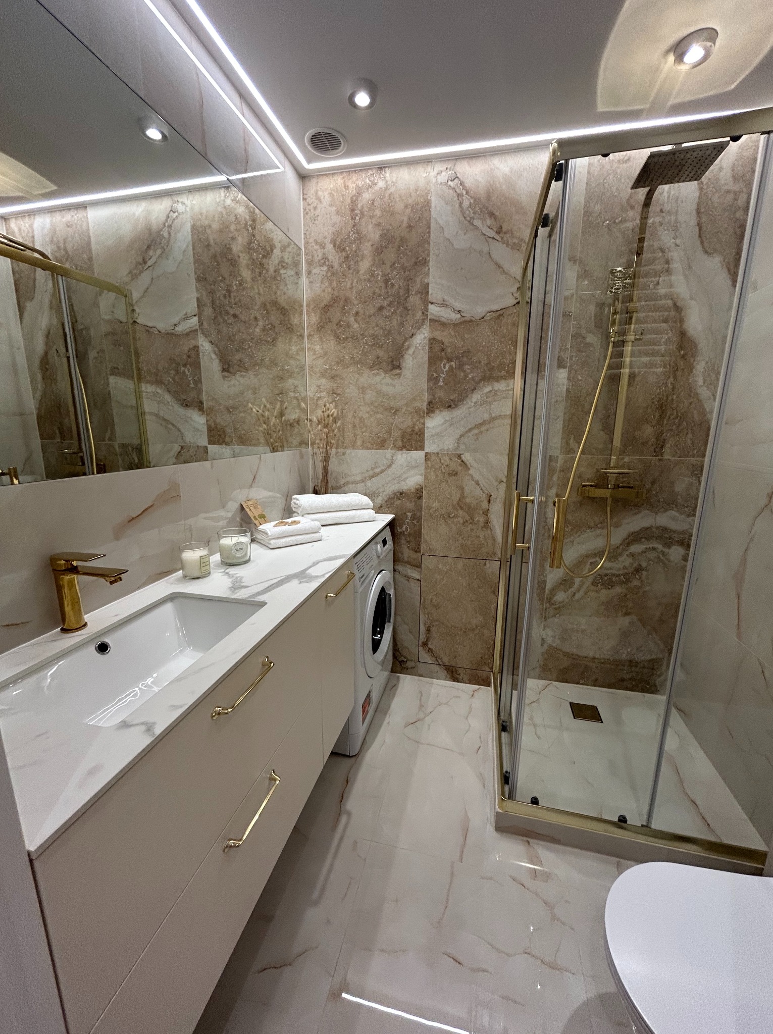 Nowoczesna łazienka z marmurowymi płytkami, złotą armaturą i kabiną prysznicową typu walk-in, oświetleniem LED oraz pralką wkomponowaną w zabudowę.