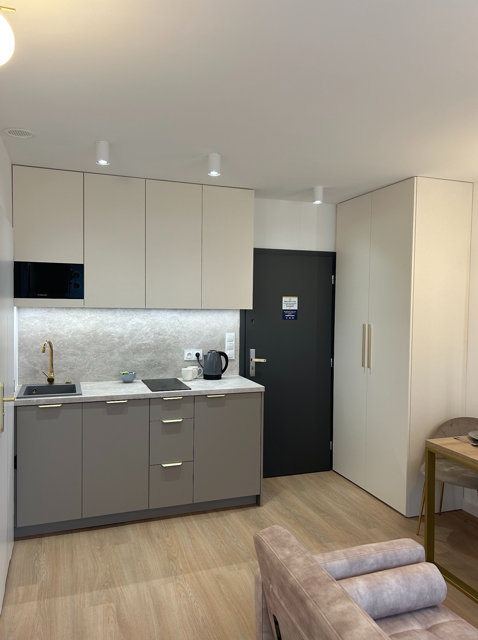 Nowoczesna, jasna kuchnia z szarymi dolnymi szafkami i beżowymi górnymi, złota bateria przy czarnym zlewie, czajnik i płyta indukcyjna na blacie, minimalistyczny design, oświetlenie punktowe.