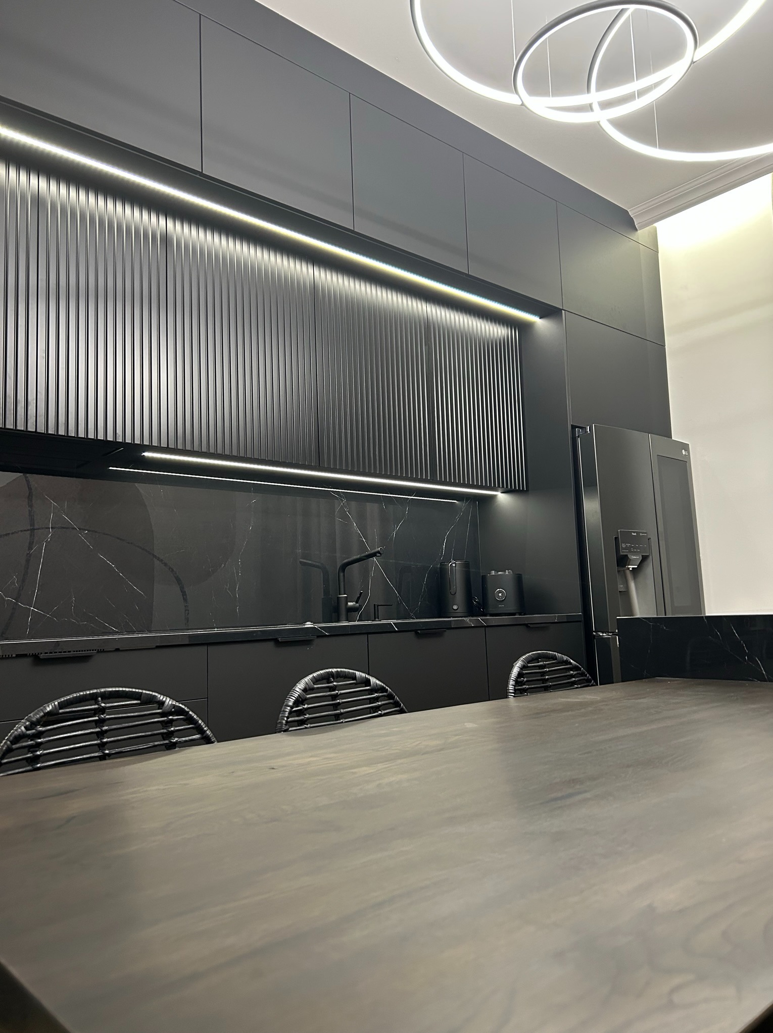 Nowoczesna, czarna kuchnia z marmurowym blatem, oświetleniem LED pod szafkami i designerską lampą sufitową, widziana zza drewnianego stołu z krzesłami o plecionych oparciach.