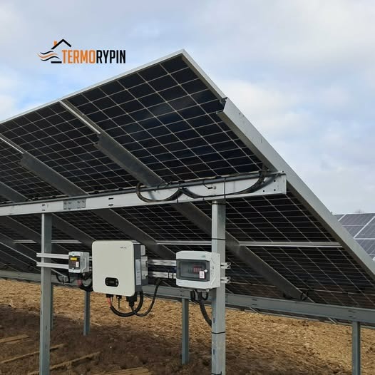 Panele słoneczne zamontowane na metalowej konstrukcji, widoczne falowniki i skrzynka elektryczna, w tle pole i pochmurne niebo.