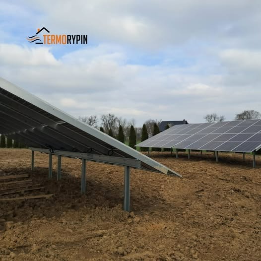 Dwa rzędy paneli słonecznych na metalowych stelażach, widok z niskiej perspektywy ukazujący konstrukcję wsporczą, z logo firmy w lewym górnym rogu i zaoranym polem w tle.