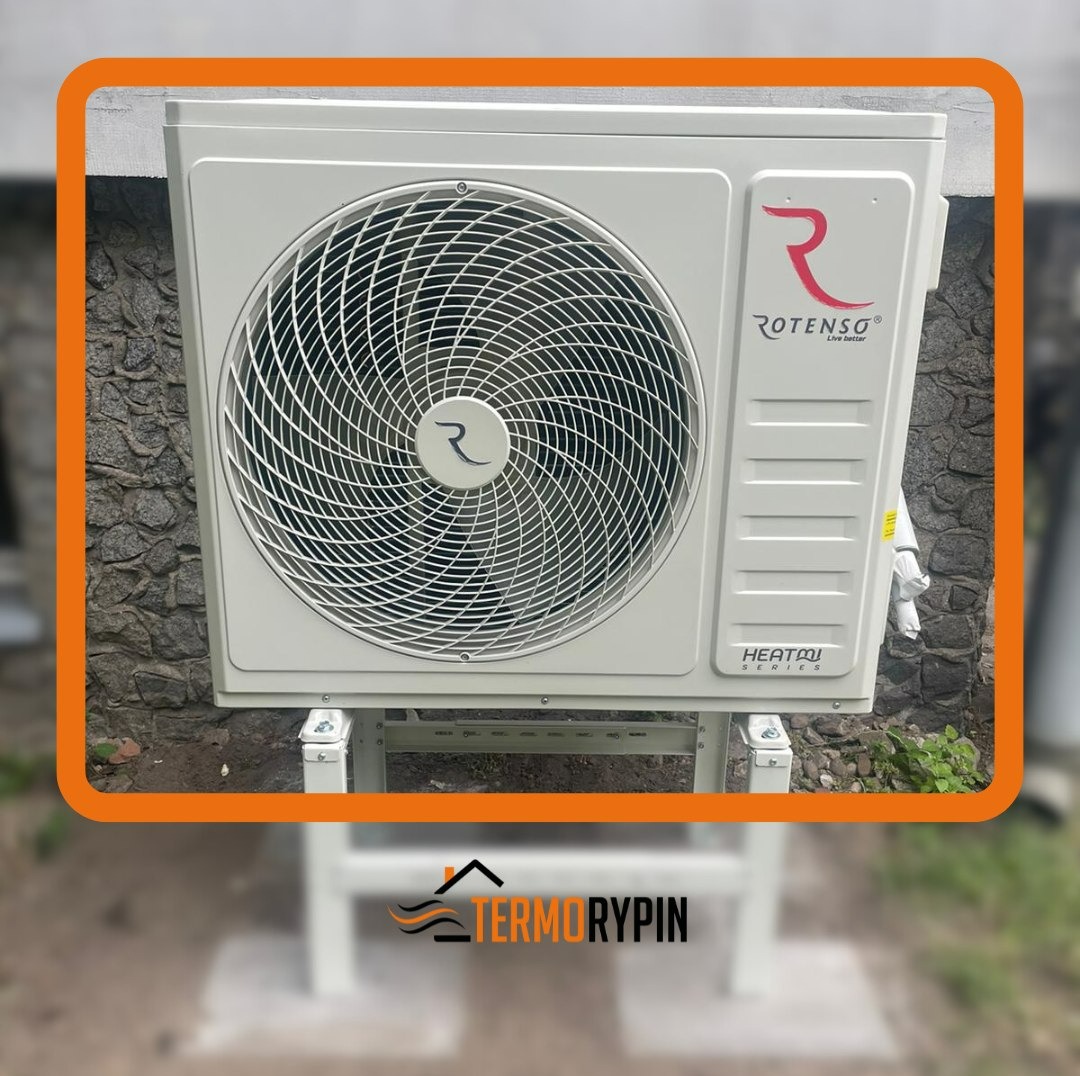 Zewnętrzna jednostka pompy ciepła Rotenso Heatpom Series zamontowana na białej konstrukcji wsporczej, z widocznym logo firmy i ścianą budynku w tle.