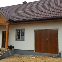 MARWOOD DOMY SZKIELETOWE SPÓŁKA Z OGRANICZONĄ ODPOWIEDZIALNOŚCIĄ - Niedokończony dom z brązowym dachem, drewnianym gankiem i garażem, widoczne narzędzia i materiały budowlane na schodach.