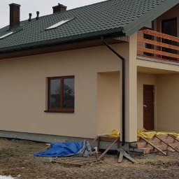 MARWOOD DOMY SZKIELETOWE SPÓŁKA Z OGRANICZONĄ ODPOWIEDZIALNOŚCIĄ - Niedawno wybudowany dom jednorodzinny z beżową elewacją, zielonym dachem, drewnianymi oknami i balkonem, widoczne prowizoryczne schody wejściowe i generator prądu.