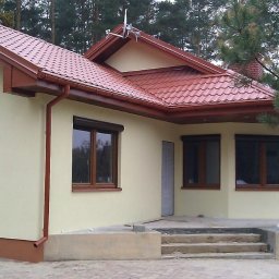 MARWOOD DOMY SZKIELETOWE SPÓŁKA Z OGRANICZONĄ ODPOWIEDZIALNOŚCIĄ - Wykończony z zewnątrz dom jednorodzinny w Legionowie z beżową elewacją, brązowymi oknami z roletami zewnętrznymi i czerwoną dachówką, otoczony drzewami.