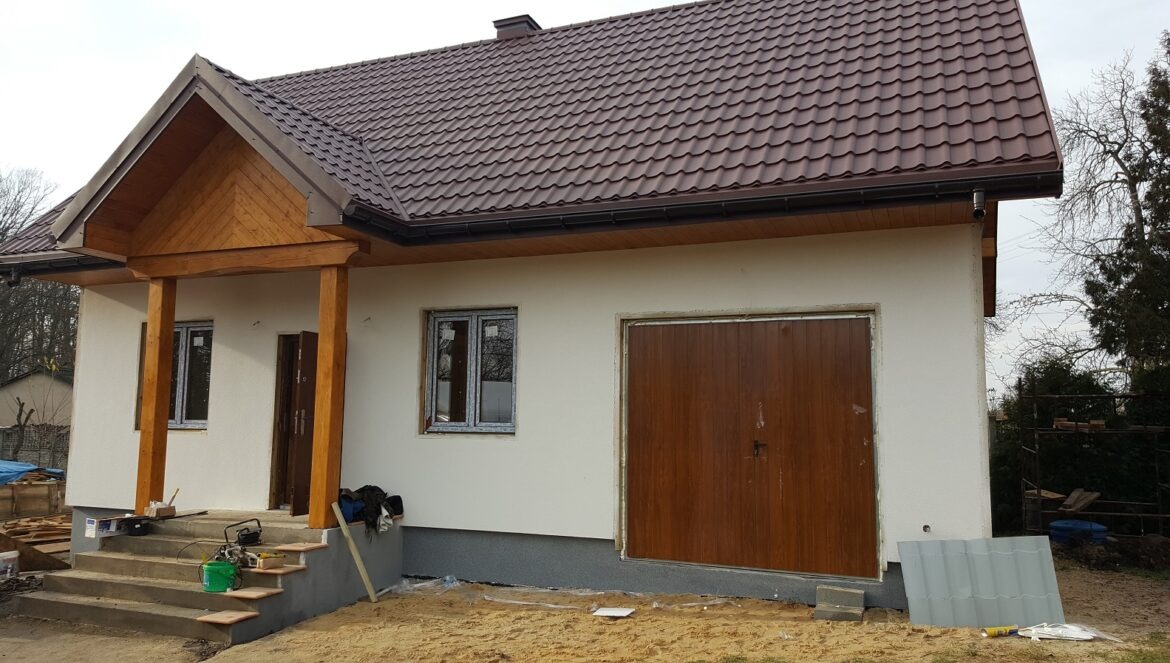 Niedokończony dom z brązowym dachem, drewnianym gankiem i garażem, widoczne narzędzia i materiały budowlane na schodach.