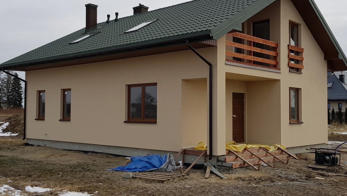 Niedawno wybudowany dom jednorodzinny z beżową elewacją, zielonym dachem, drewnianymi oknami i balkonem, widoczne prowizoryczne schody wejściowe i generator prądu.
