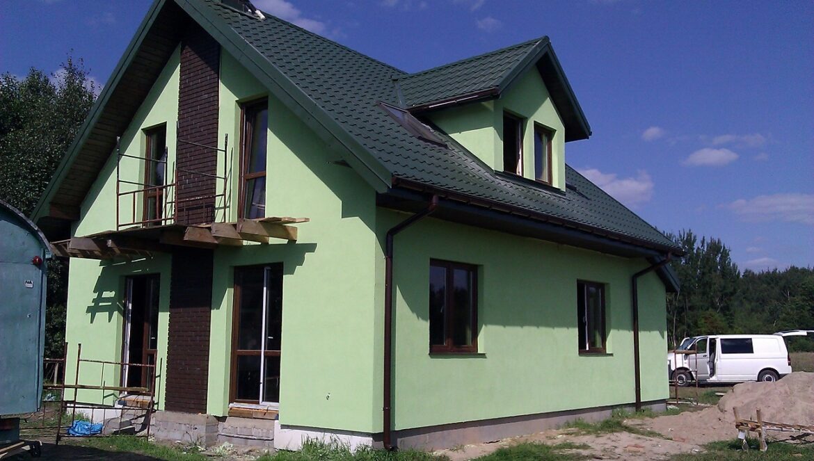 Jasnozielony dom szkieletowy z ciemnozielonym dachem, widoczne rusztowania i fragment elewacji z cegły, w tle las i biały bus.
