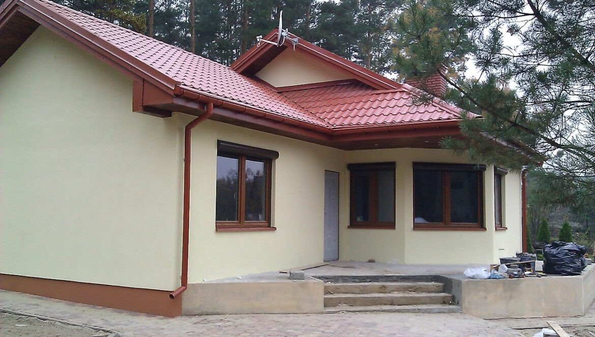 Wykończony z zewnątrz dom jednorodzinny w Legionowie z beżową elewacją, brązowymi oknami z roletami zewnętrznymi i czerwoną dachówką, otoczony drzewami.