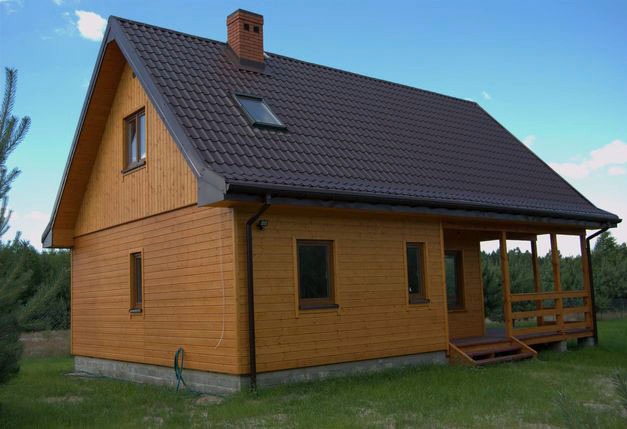 Gotowy, parterowy dom z drewnianą elewacją, brązowym dachem z dachówką i małą werandą z drewnianą balustradą.