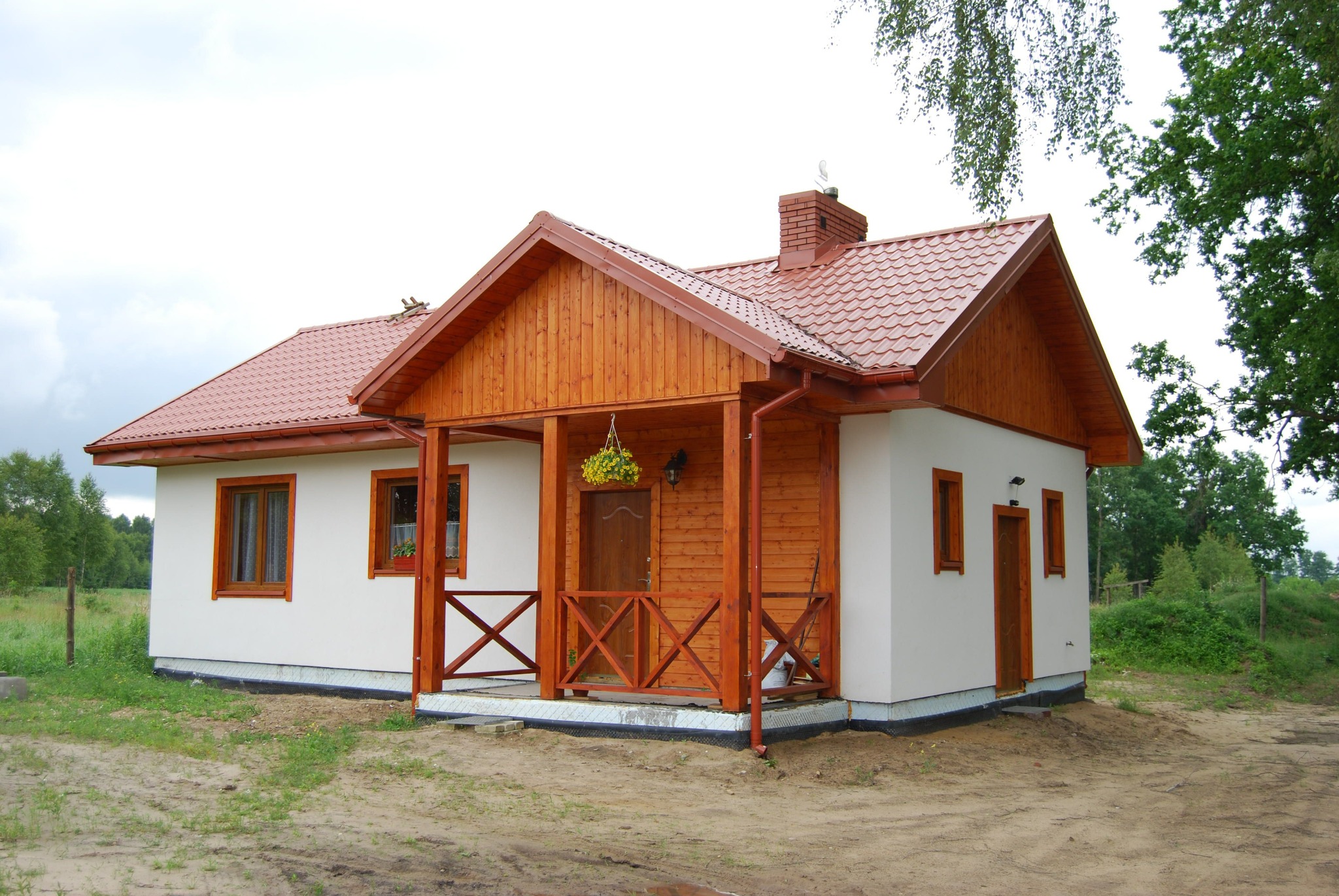 Niewielki parterowy dom z drewnianą werandą, czerwoną dachówką i białymi ścianami, widok z przodu na tle zieleni.