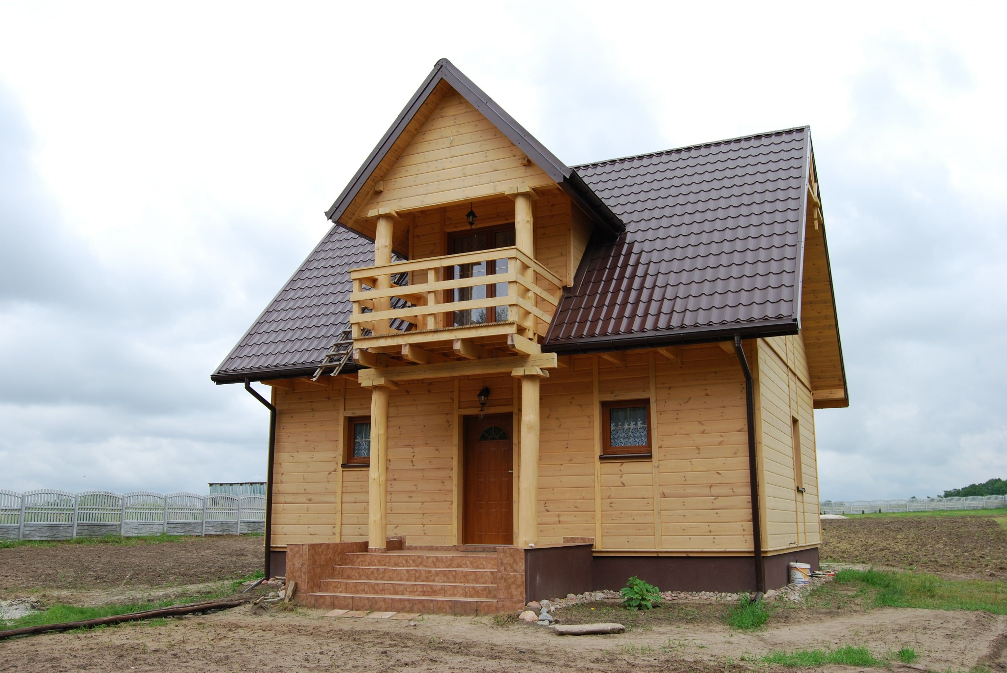 Dwukondygnacyjny dom z jasnego drewna z balkonem, brązowym dachem i schodami wejściowymi z terakoty, widok z przodu na tle zaoranego pola i ogrodzenia.