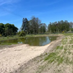Zbiornik wodny z piaszczystym brzegiem i trawą, widok z góry, błękitne niebo, drzewa w tle, częściowo widoczny budynek z białej cegły po prawej.