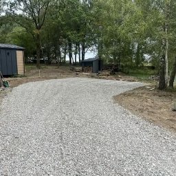 Paweł Zalewski usługi budowlane - Wysypany żwirem podjazd do domku letniskowego z szopą na drewno w tle, otoczony zielenią. Widoczne narzędzia ogrodnicze. Kadr z poziomu gruntu.