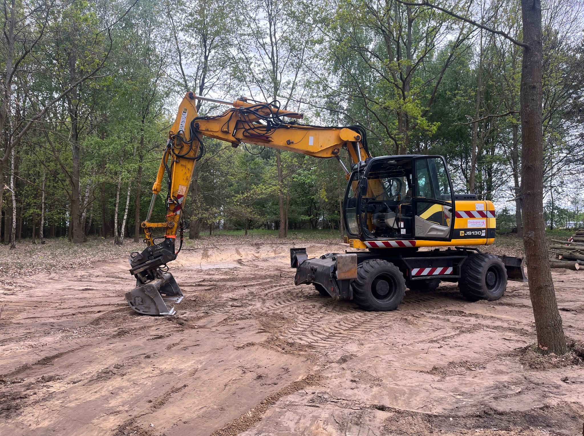 Żółta koparka kołowa JCB JS130D na placu budowy z wykopem, w tle drzewa. Widoczne ślady opon na ziemi i ścięte pnie drzew. Maszyna przygotowana do pracy.