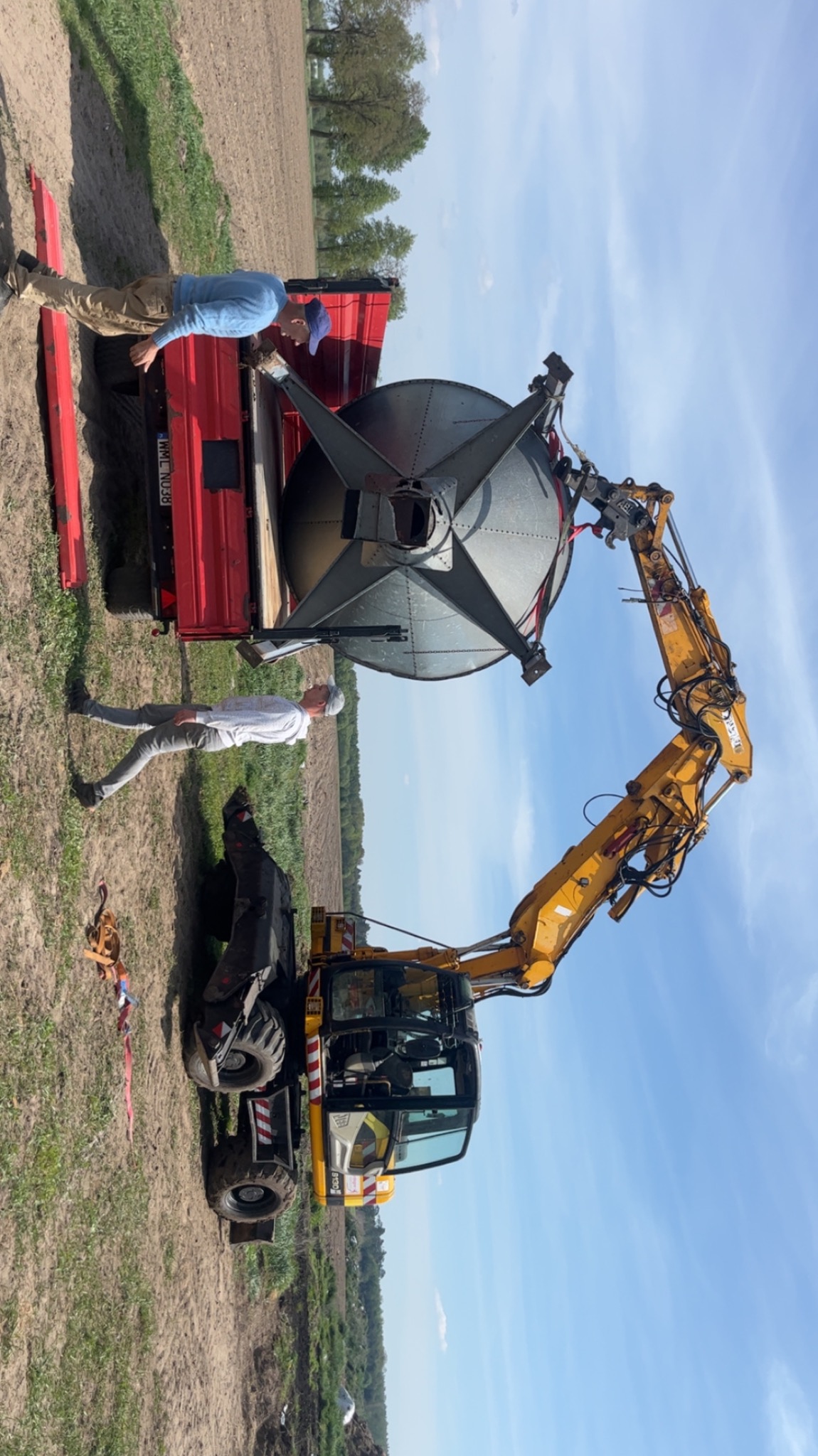 Żółta koparka transportuje metalowy zbiornik na przyczepie, nadzorowana przez dwóch mężczyzn na tle pola i błękitnego nieba.