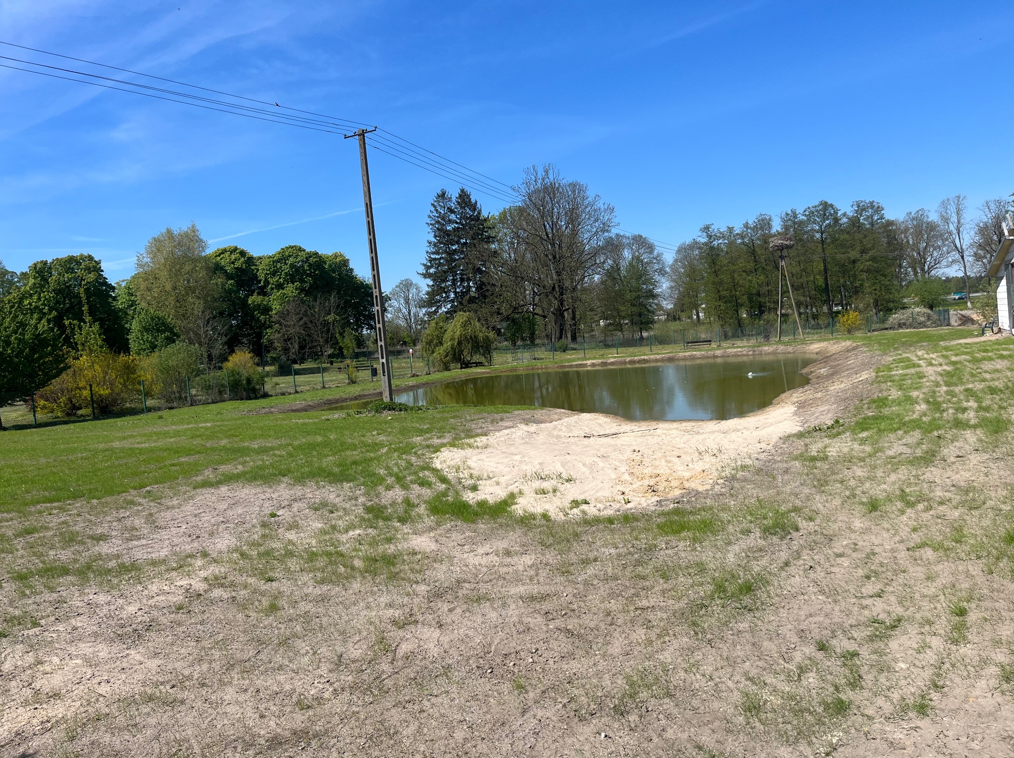 Wyrobisko ziemne z częściowo wypełnionym zbiornikiem wodnym, otoczone zielenią i drzewami, w słoneczny dzień. Widoczny słup energetyczny i gniazdo bocianie.