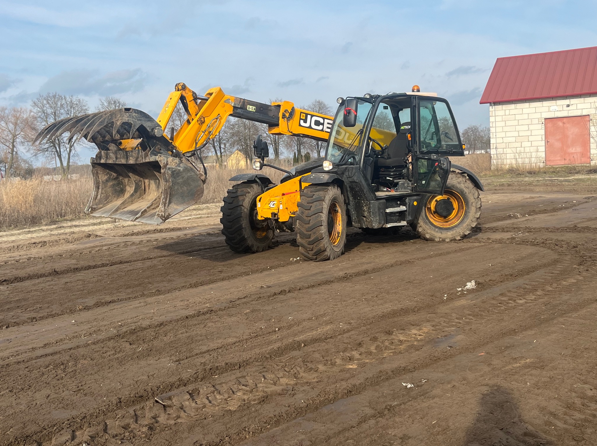 Żółto-czarna ładowarka teleskopowa JCB z łyżką i chwytakiem na polu. W tle budynek z czerwonym dachem i drzewa. Ślady opon na ziemi.