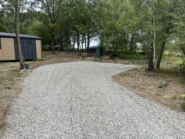 Wysypany żwirem podjazd do domku letniskowego z szopą na drewno w tle, otoczony zielenią. Widoczne narzędzia ogrodnicze. Kadr z poziomu gruntu.