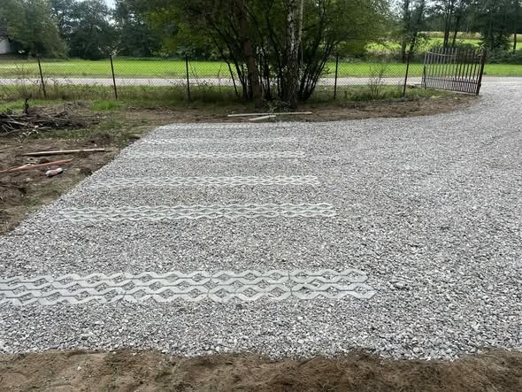 Żwirowa nawierzchnia z ażurowymi płytami betonowymi, tworzącymi wzór. W tle widoczne ogrodzenie i zieleń. Ujęcie z poziomu gruntu.