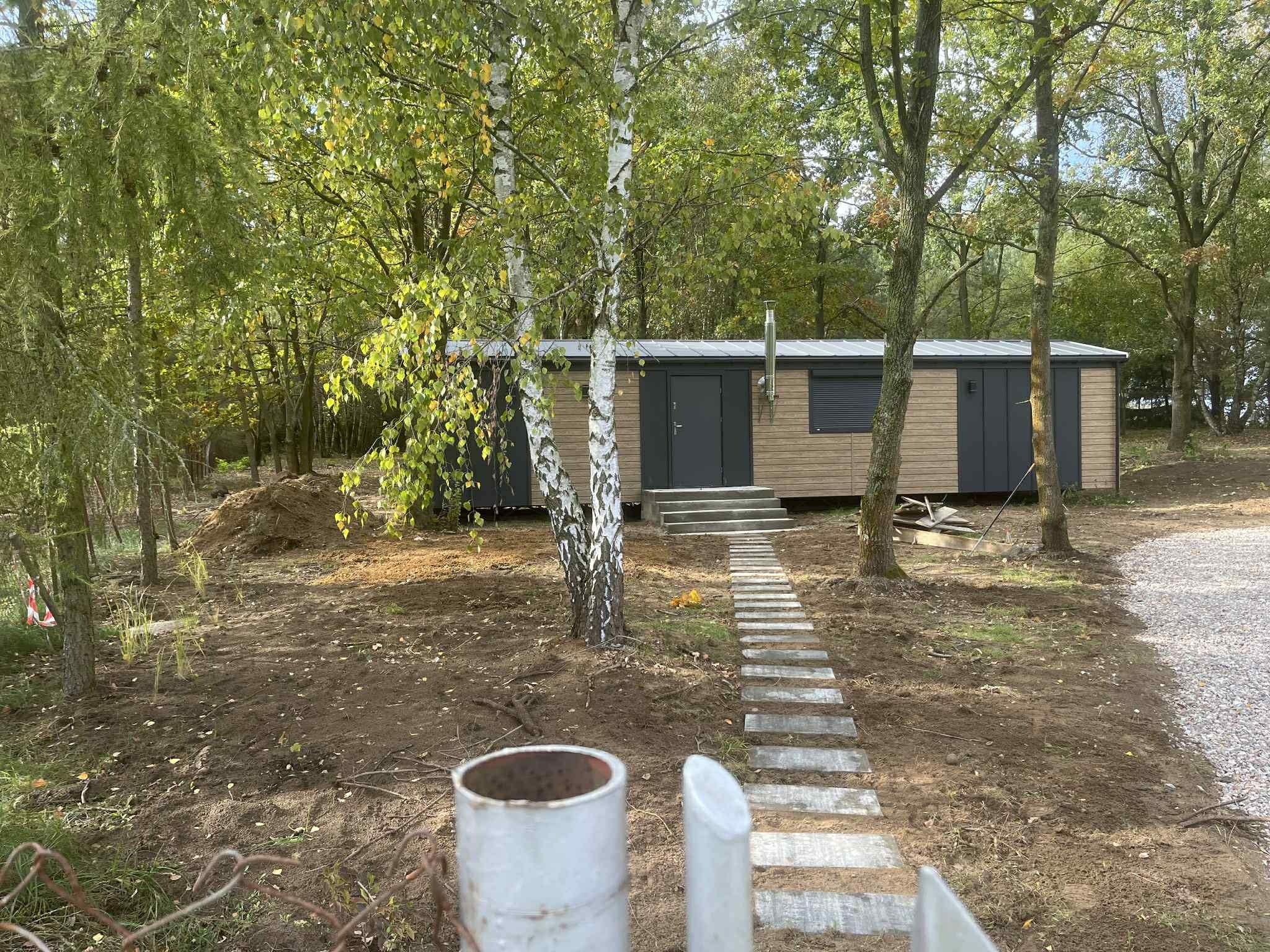 Nowoczesny, drewniany domek letniskowy z szarymi elementami, otoczony drzewami. Ścieżka z płyt prowadzi do wejścia. Widoczne ślady prac ziemnych i fragment ogrodzenia.