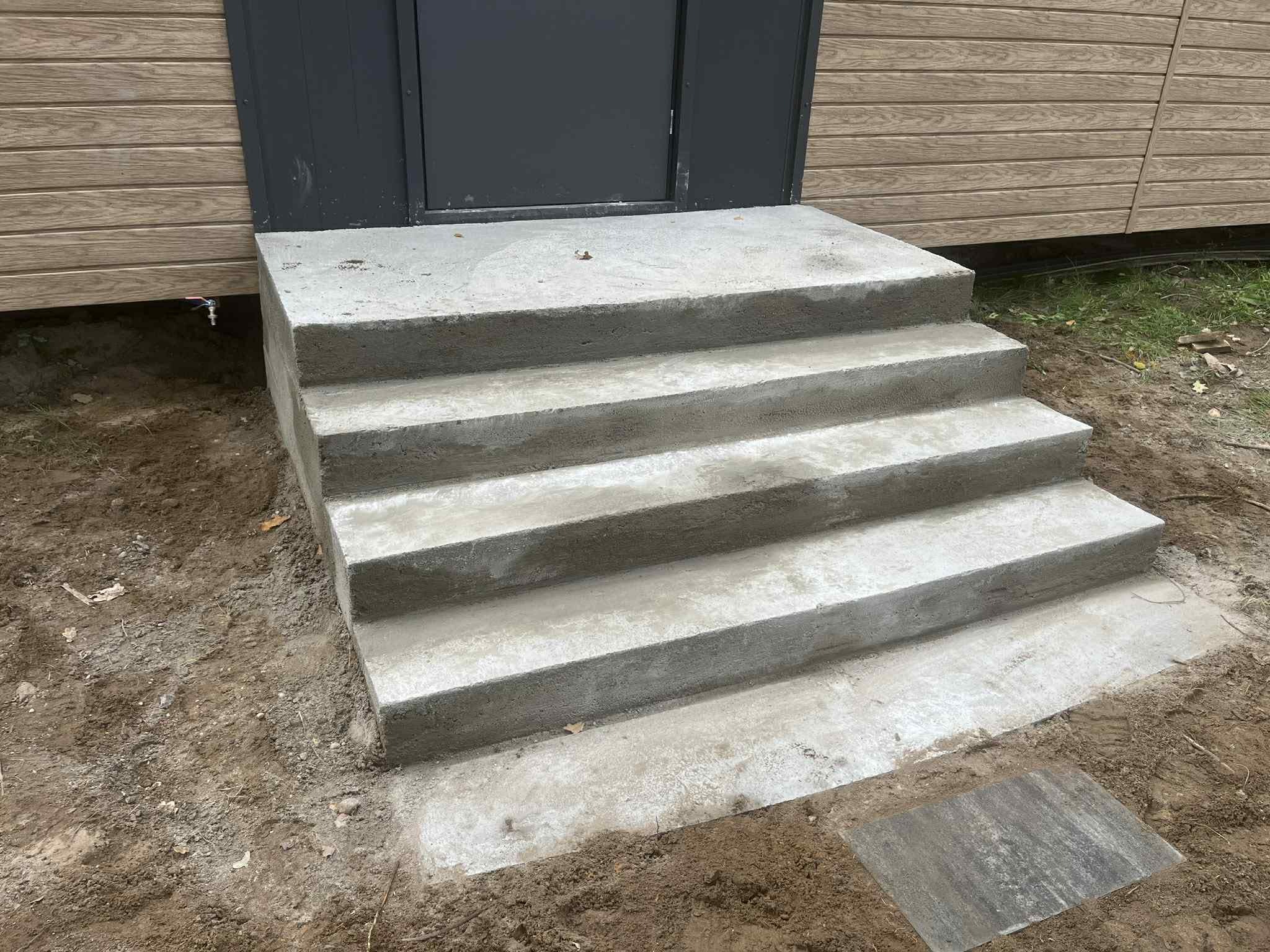 Betonowe schody wejściowe do budynku z drewnianą elewacją. Czterostopniowe schody prowadzą do ciemnych drzwi. U dołu widoczna kostka brukowa i ziemia.