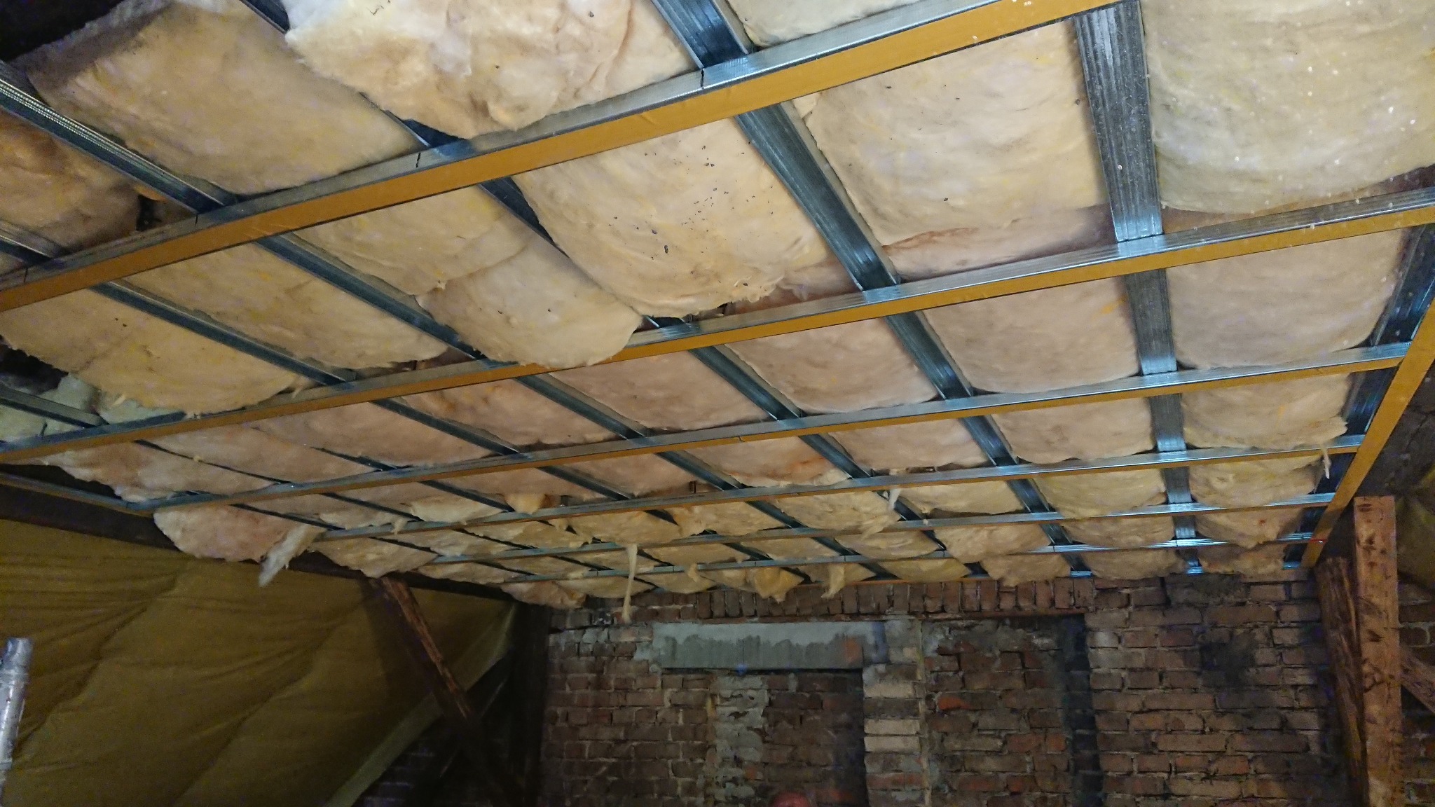 Ocieplenie poddasza wełną mineralną, widoczne metalowe profile konstrukcyjne. Prace wykończeniowe w surowym wnętrzu z ceglaną ścianą i widoczną izolacją.