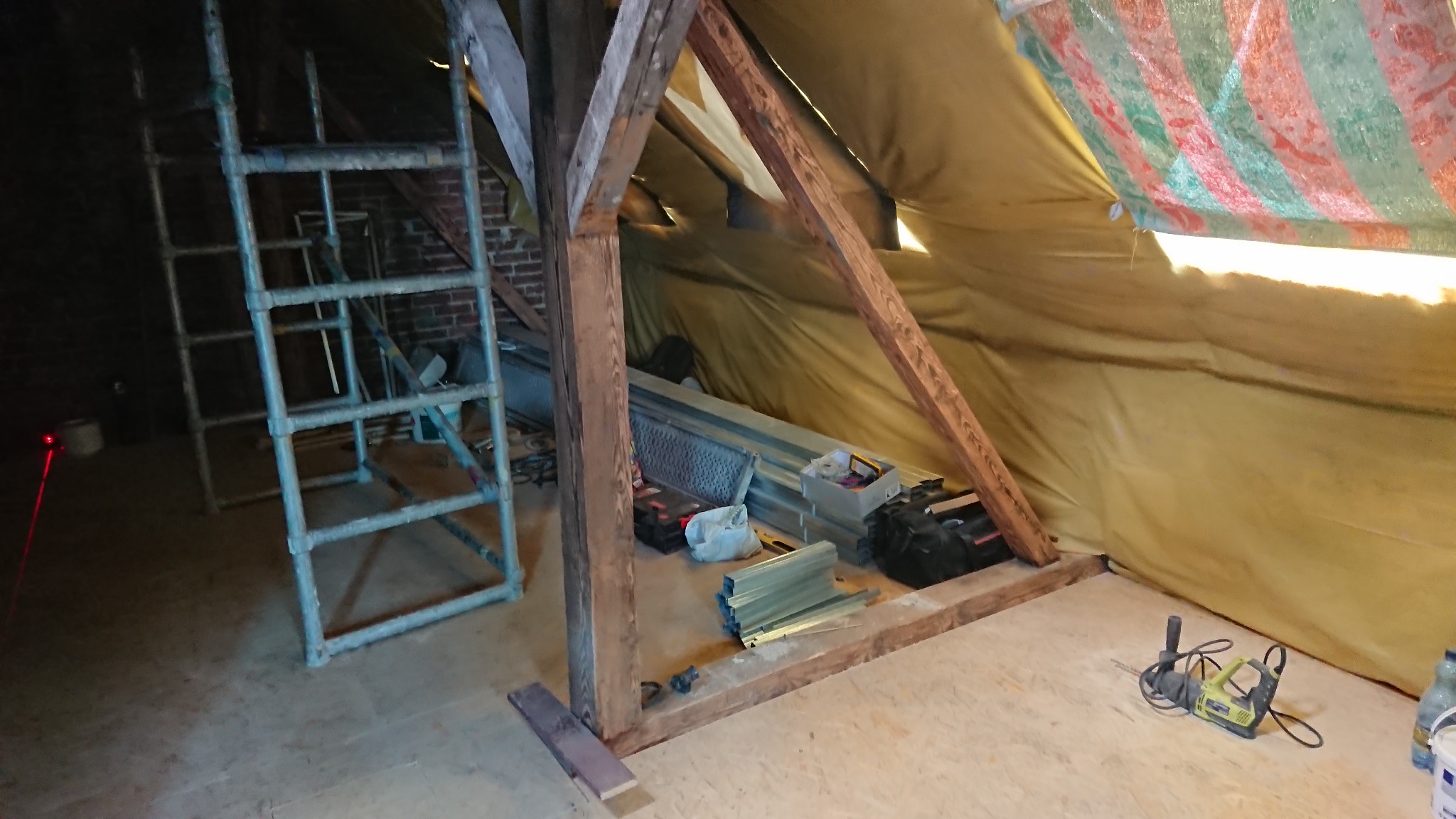 Wnętrze poddasza w trakcie prac dekarskich: widoczna więźba, rusztowanie, narzędzia i materiały budowlane. Oświetlenie robocze, folia ochronna na dachu.