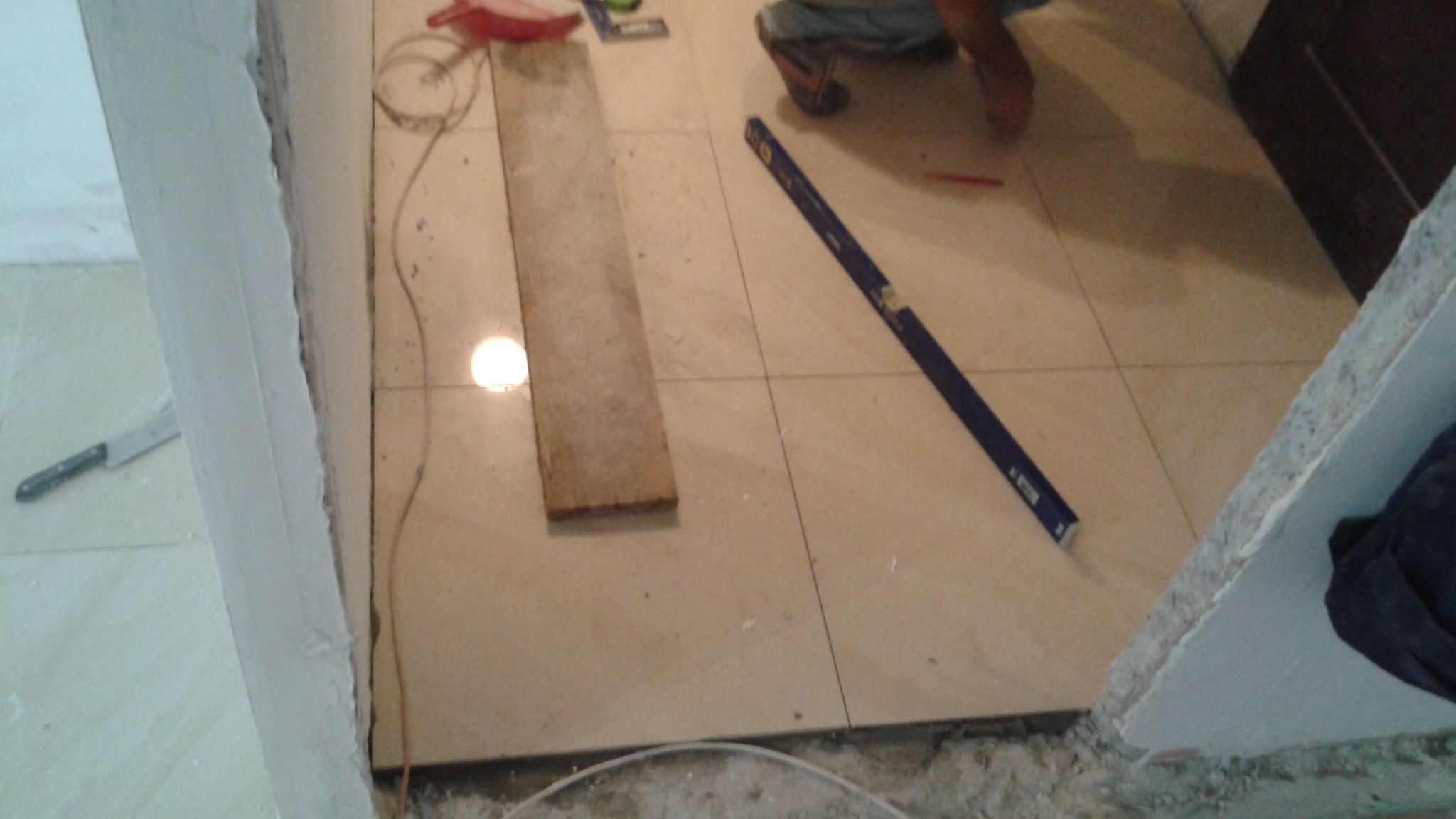 Układanie płytek podłogowych: poziomica, deska i narzędzia na beżowych płytkach. Widoczne fragmenty ścian w trakcie prac wykończeniowych.