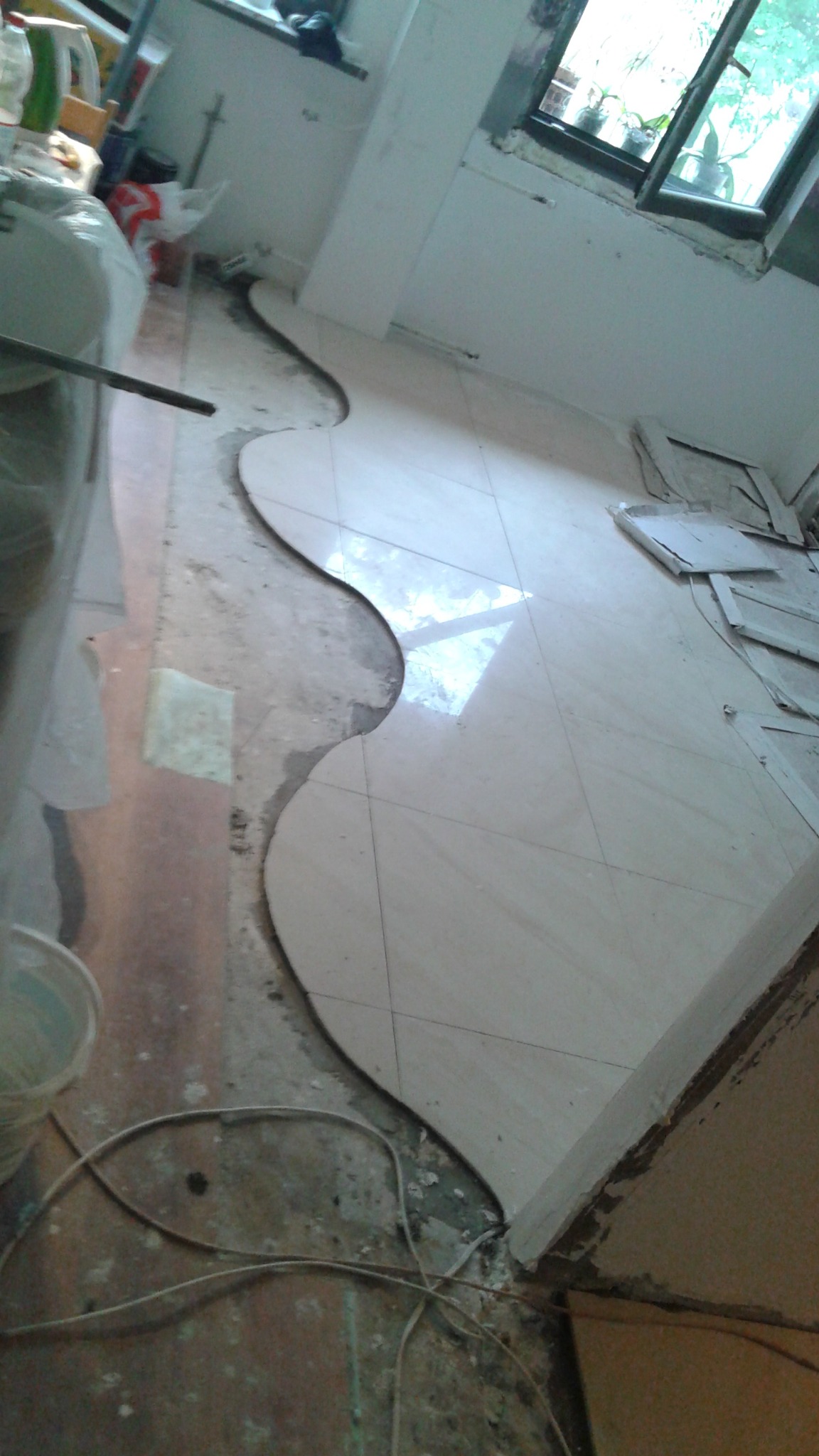 Układanie płytek podłogowych w pomieszczeniu z nieregularnym kształtem, widoczne narzędzia i materiały budowlane. Częściowo ułożona podłoga z jasnych płytek.