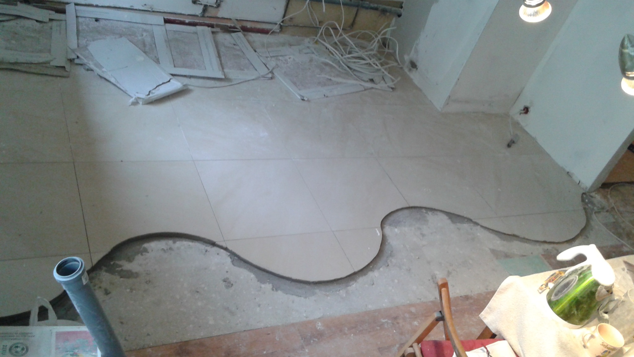 Ułożone jasne płytki podłogowe z wyciętym, nieregularnym kształtem, widoczne kable i resztki materiałów budowlanych. Fragment rury kanalizacyjnej w lewym dolnym rogu.