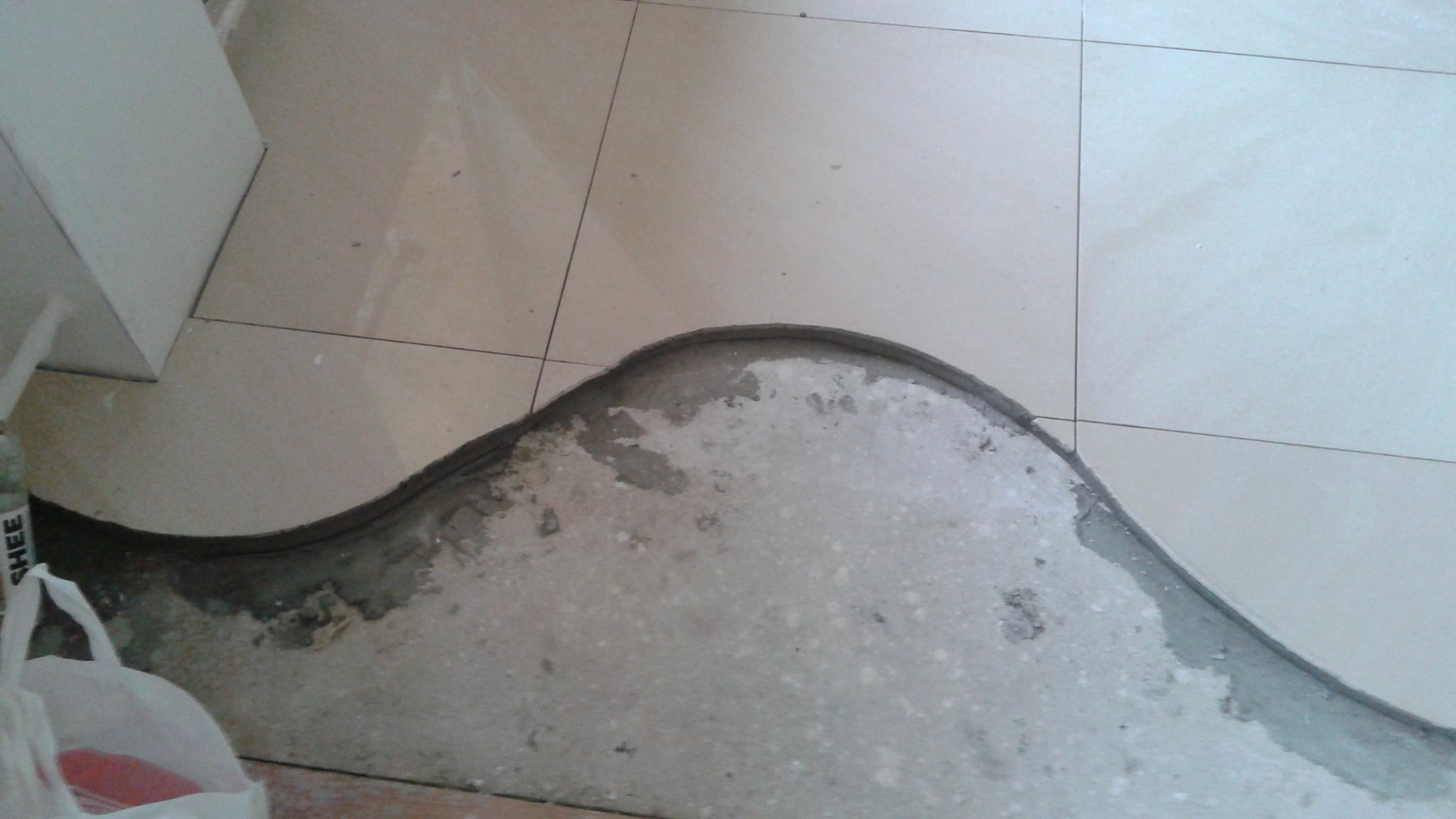 Układanie płytek podłogowych w łazience, widoczny fragment podłogi z wykończonym brzegiem, jasne płytki i szara zaprawa. Nietypowe wykończenie podłogi.