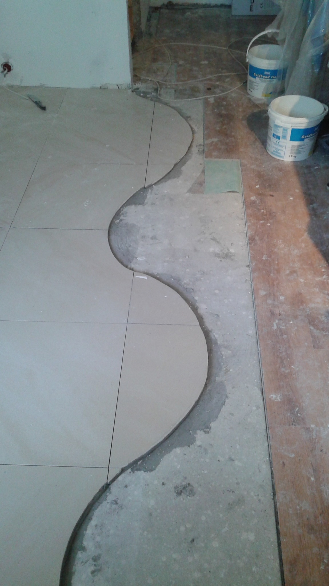 Układanie płytek podłogowych z wycięciami w kształcie fal, widoczne narzędzia i materiały budowlane. Prace wykończeniowe w trakcie realizacji.