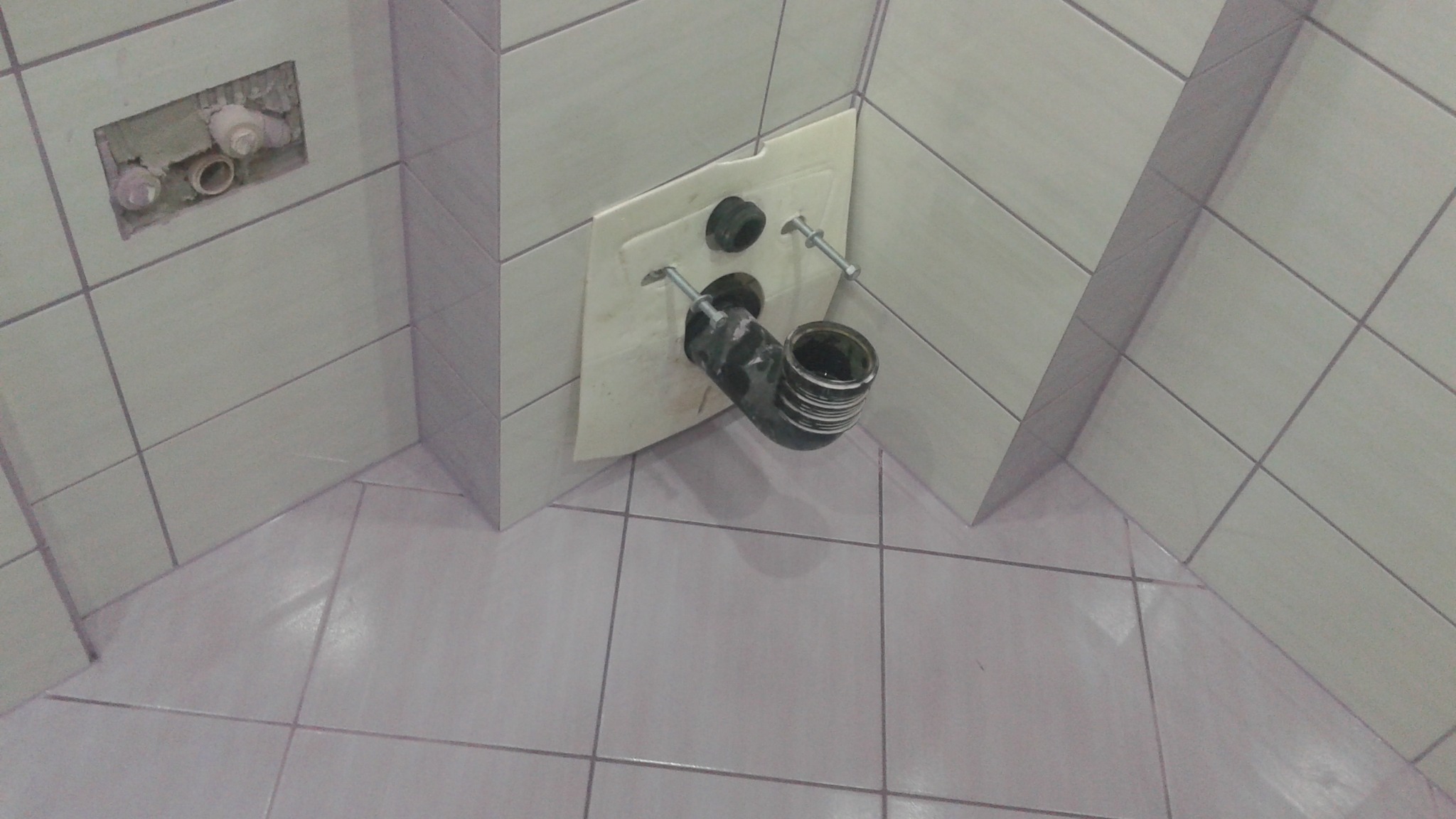 Zamontowana rura odpływowa w łazience z jasnymi płytkami. Widoczne mocowania i fragment instalacji w ścianie. Ujęcie z góry, blisko podłogi.