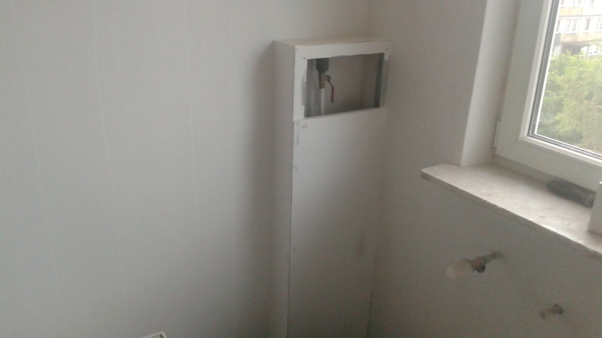 Biała zabudowa pionu wodno-kanalizacyjnego z otwartym fragmentem, widoczne rury i zawór. Obok okno z parapetem. Ściany w jasnym odcieniu. Ujęcie z dołu.