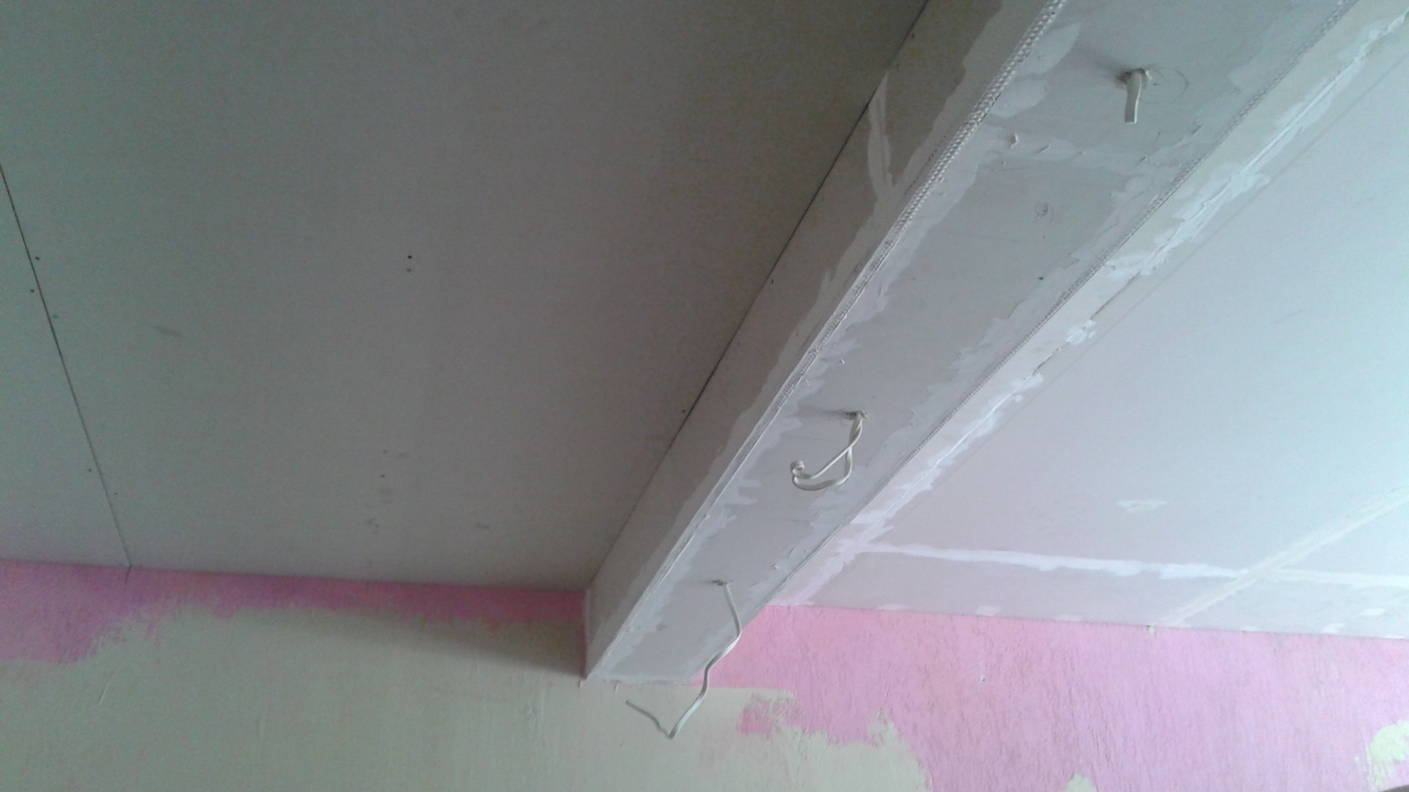 Sufit podwieszany z płyt gipsowo-kartonowych w trakcie montażu, widoczne profile i łączenia. Fragment ściany z resztkami farby. Ujęcie z dołu, perspektywa z narożnika.