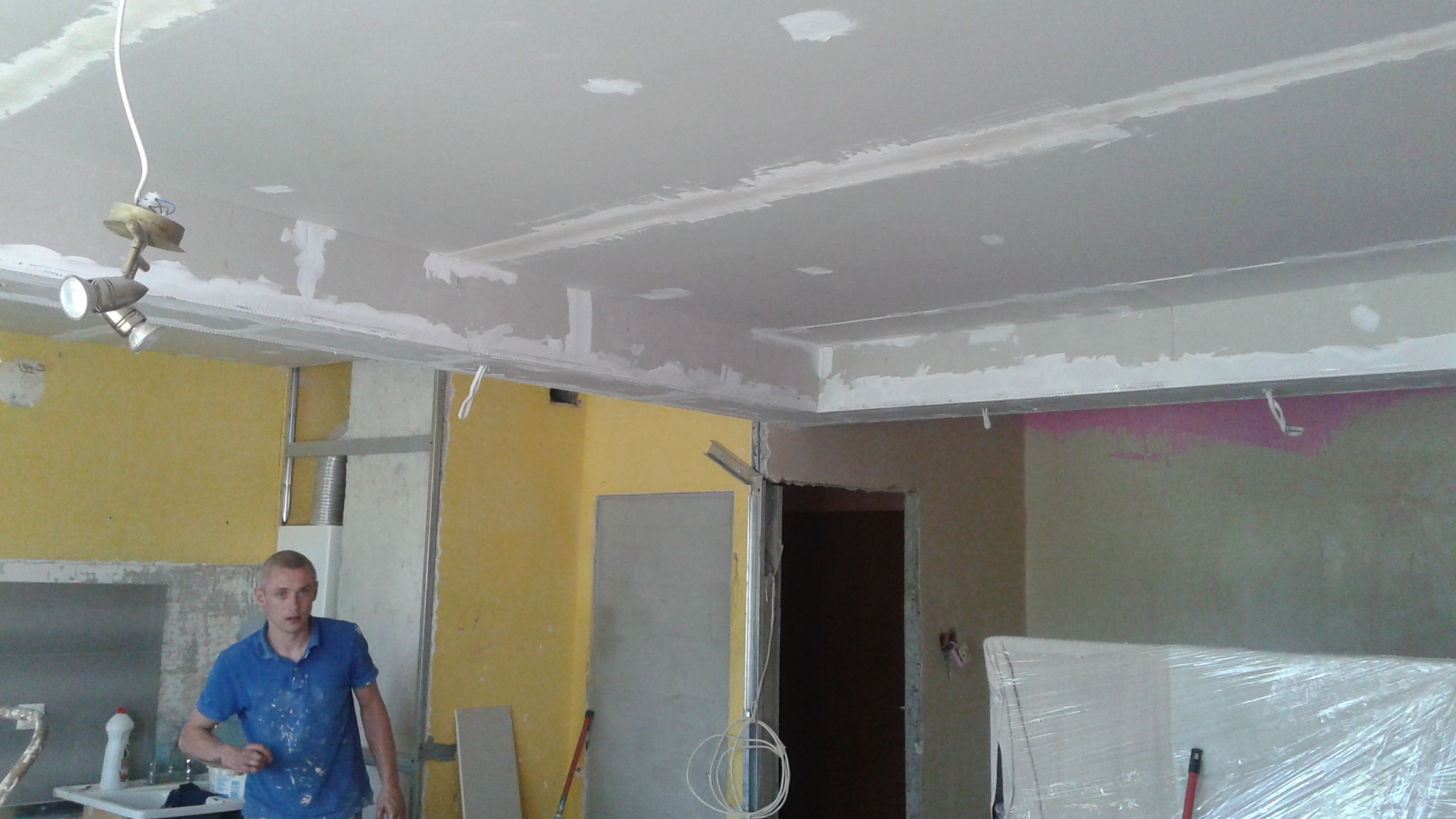 Montaż sufitu podwieszanego z płyt gipsowo-kartonowych w pomieszczeniu z żółtymi ścianami. Widoczny pracownik w niebieskiej koszulce. Prace wykończeniowe w toku.
