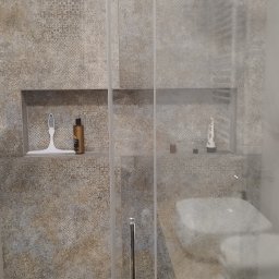 AlexBud - Kabina prysznicowa z przesuwnymi drzwiami, wyłożona wzorzystymi płytkami w odcieniach beżu i szarości, z wnękami na kosmetyki i czarnym dywanikiem z geometrycznym wzorem na podłodze.