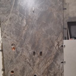 AlexBud - Ściana pokryta szarą płytką imitującą marmur, z widocznymi otworami na instalacje i elementami mocującymi, ukazująca etap prac wykończeniowych w pomieszczeniu.