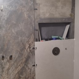 AlexBud - Montaż płytek imitujących marmur w łazience, widoczne krzyżyki dystansowe i przygotowane miejsce na półkę w ścianie, podłoga zabezpieczona folią.