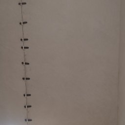 AlexBud - Ściana w trakcie układania płytek, widoczne krzyżyki dystansowe, narzędzia glazurnicze i zabezpieczona folią podłoga.