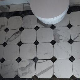 AlexBud - Ułożona podłoga w łazience z białych, ośmiokątnych płytek imitujących marmur, z czarnymi kwadratami w narożnikach i białą miską toaletową w tle, częściowo zakryta folią ochronną.