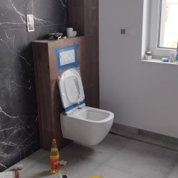 AlexBud - Podwieszana miska WC w trakcie montażu w łazience z marmurową ścianą, widoczne narzędzia i butelka na podłodze.
