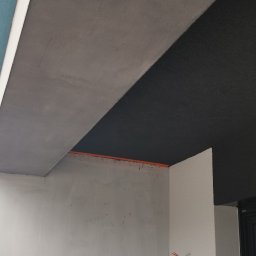 AlexBud - Narożnik pomieszczenia w trakcie prac wykończeniowych: szara gładź sufitowa, surowa ściana przygotowana pod malowanie, wystające przewody elektryczne.