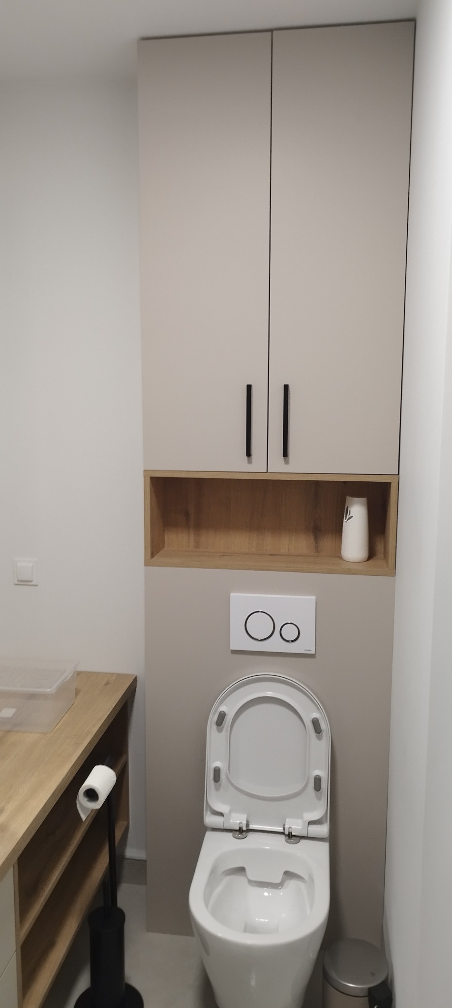 Nowoczesna toaleta z otwartą deską sedesową, zabudowana szafka wisząca z półką na dekoracyjny wazon, przycisk spłuczki z dwoma okręgami, obok szafka z blatem i czarny stojak na papier toaletowy...