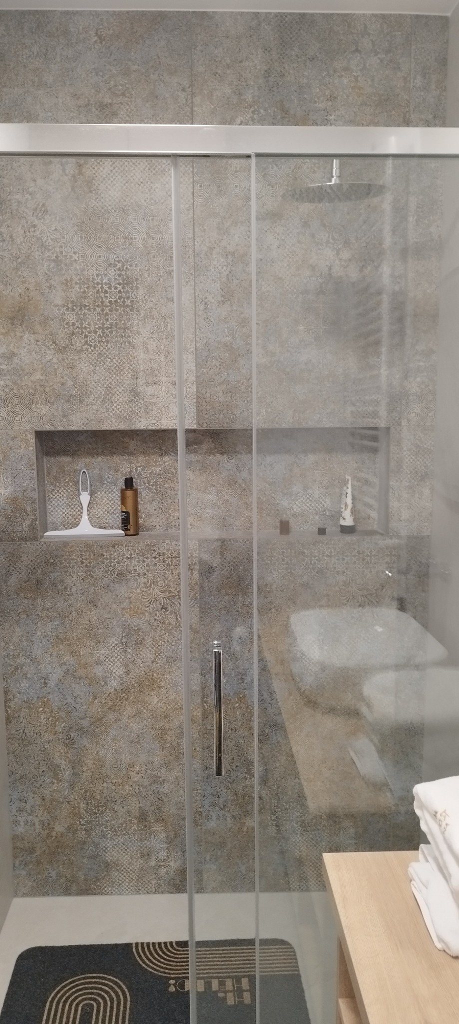 Kabina prysznicowa z przesuwnymi drzwiami, wyłożona wzorzystymi płytkami w odcieniach beżu i szarości, z wnękami na kosmetyki i czarnym dywanikiem z geometrycznym wzorem na podłodze.