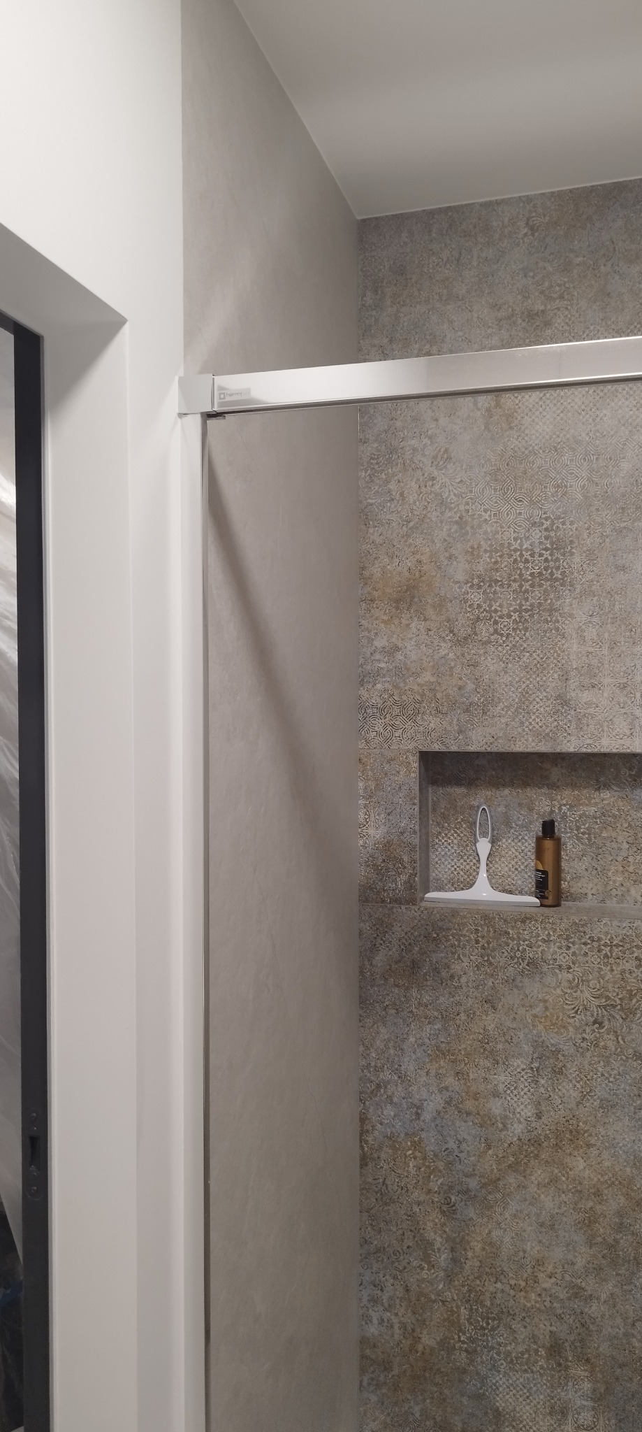 Szklana kabina prysznicowa z chromowanymi profilami, wbudowana półka z akcesoriami do pielęgnacji, ściany wyłożone płytkami w odcieniach szarości i brązu z delikatnym wzorem.