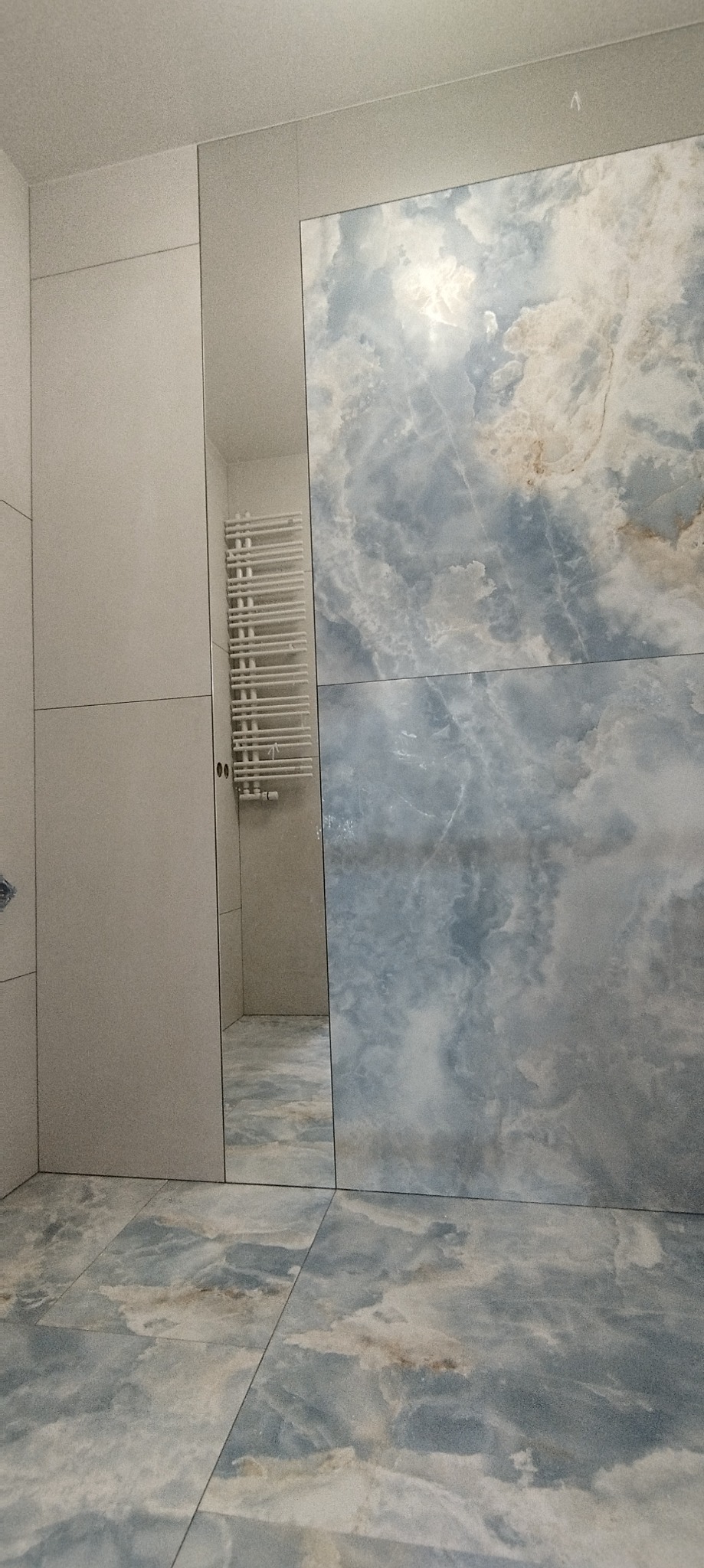 Wykończona łazienka z płytkami imitującymi marmur w odcieniach błękitu i szarości, widoczny grzejnik drabinkowy w wąskiej wnęce.