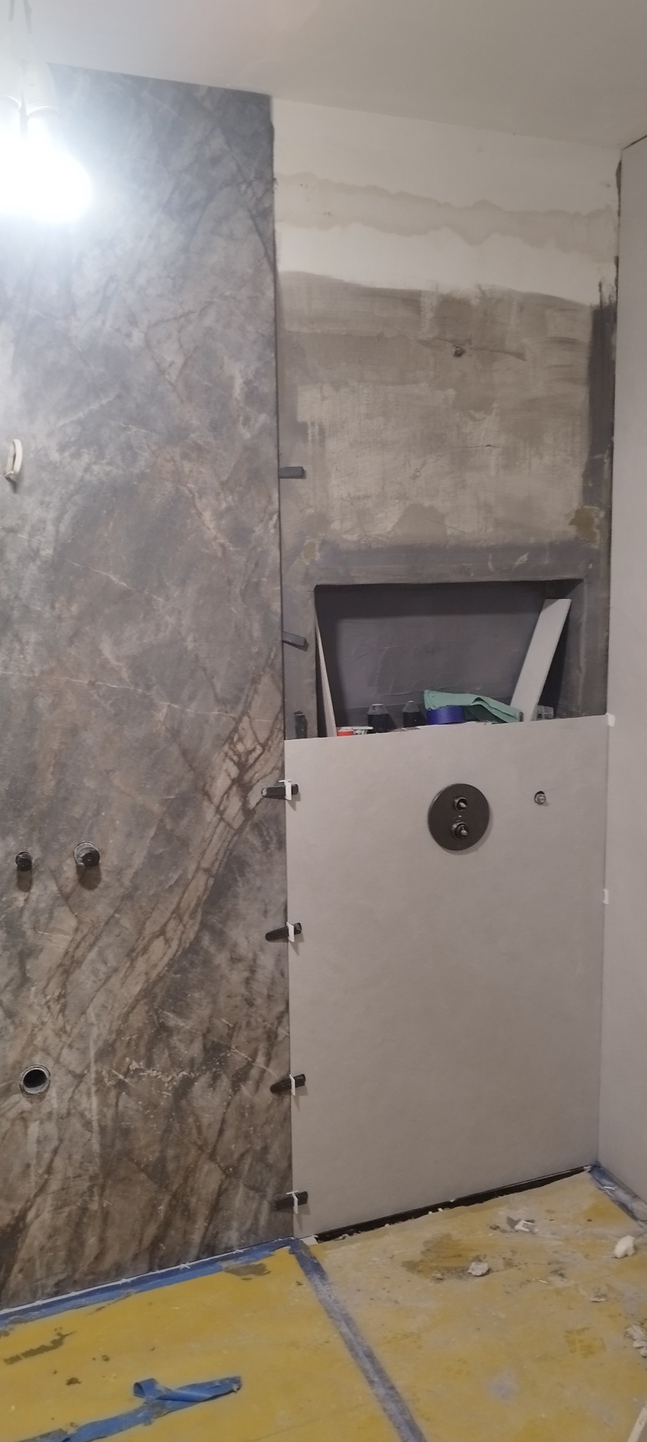 Montaż płytek imitujących marmur w łazience, widoczne krzyżyki dystansowe i przygotowane miejsce na półkę w ścianie, podłoga zabezpieczona folią.